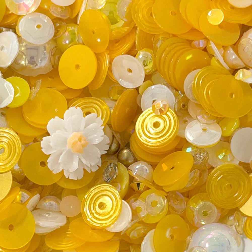 A close-up pile of craft embellishments, including many translucent yellow and white round sequins, spiral disc beads, clear faceted beads, and a small white flower-shaped bead.