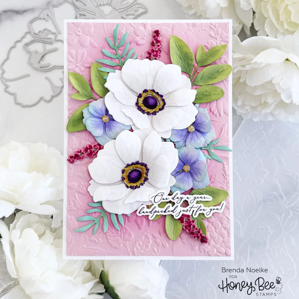 A handmade layered card featuring three white anemone flowers with purple centers, blue-purple blossoms, green leaves, and pink background embossing, styled with white flowers and metal dies. Text at the bottom reads Brenda Noeike for Honey Bee Stamper.