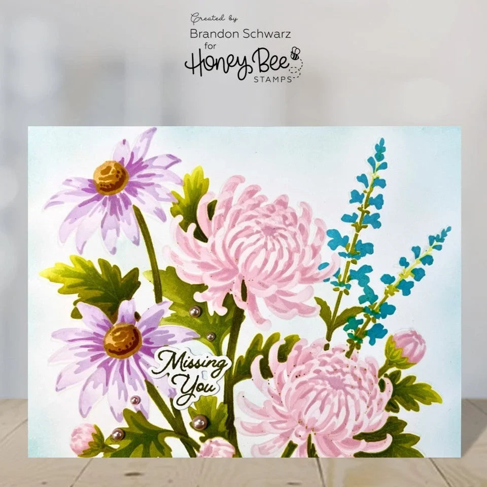 A floral greeting card featuring pink chrysanthemums, purple daisies, blue wildflowers, green leaves, and the words 'Missing You' in script. The card is displayed upright on a light wood surface against a soft gray background, with 'Created by Brandon Schwarz for Honey Bee STAMPS' printed above.