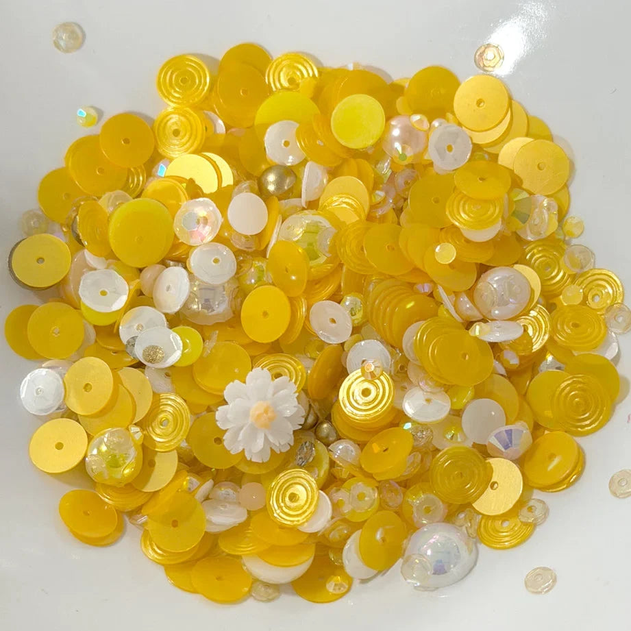 A pile of yellow and white craft sequins, beads, and small decorative pieces on a plain white background.