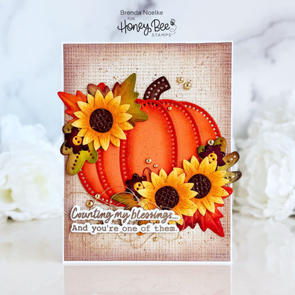 A handmade autumn-themed greeting card featuring a layered orange pumpkin, yellow sunflowers, fall leaves, and bumblebees on a burlap-textured background. The card is displayed upright on a white surface with soft white flowers blurred in the background.
