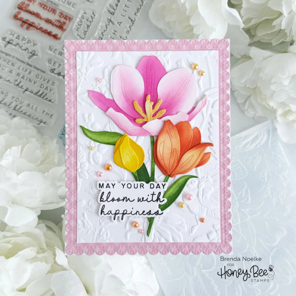 A handmade floral greeting card with embossed white background, a pink scalloped border, and layered paper flowers in pink, orange, and yellow. The card reads “MAY YOUR DAY bloom with happiness” and is photographed among white flowers, with Honey Bee Stamp branding visible.