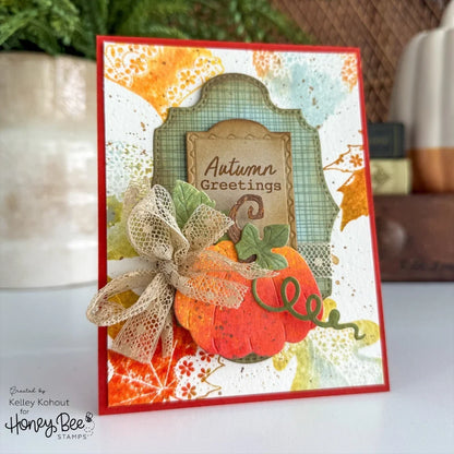 A handmade autumn-themed greeting card with a red border, layered paper arches, a pumpkin embellishment, and the words 'Autumn Greetings' on a beige label. The card is photographed on a white surface with a blurred background containing a plant and decorative objects.