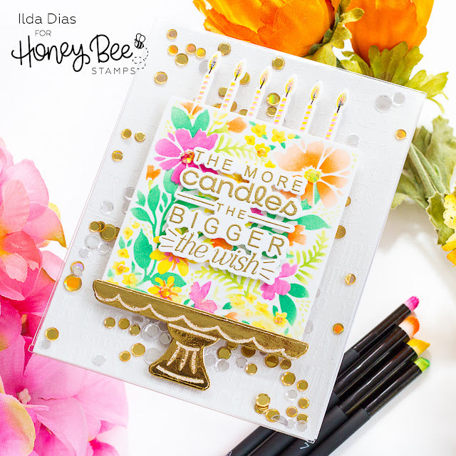 A handmade greeting card featuring a colorful birthday cake illustration with candles and the text 'THE MORE candles THE BIGGER the wish'. The card is styled with gold confetti, surrounded by flowers and craft tools on a white background.